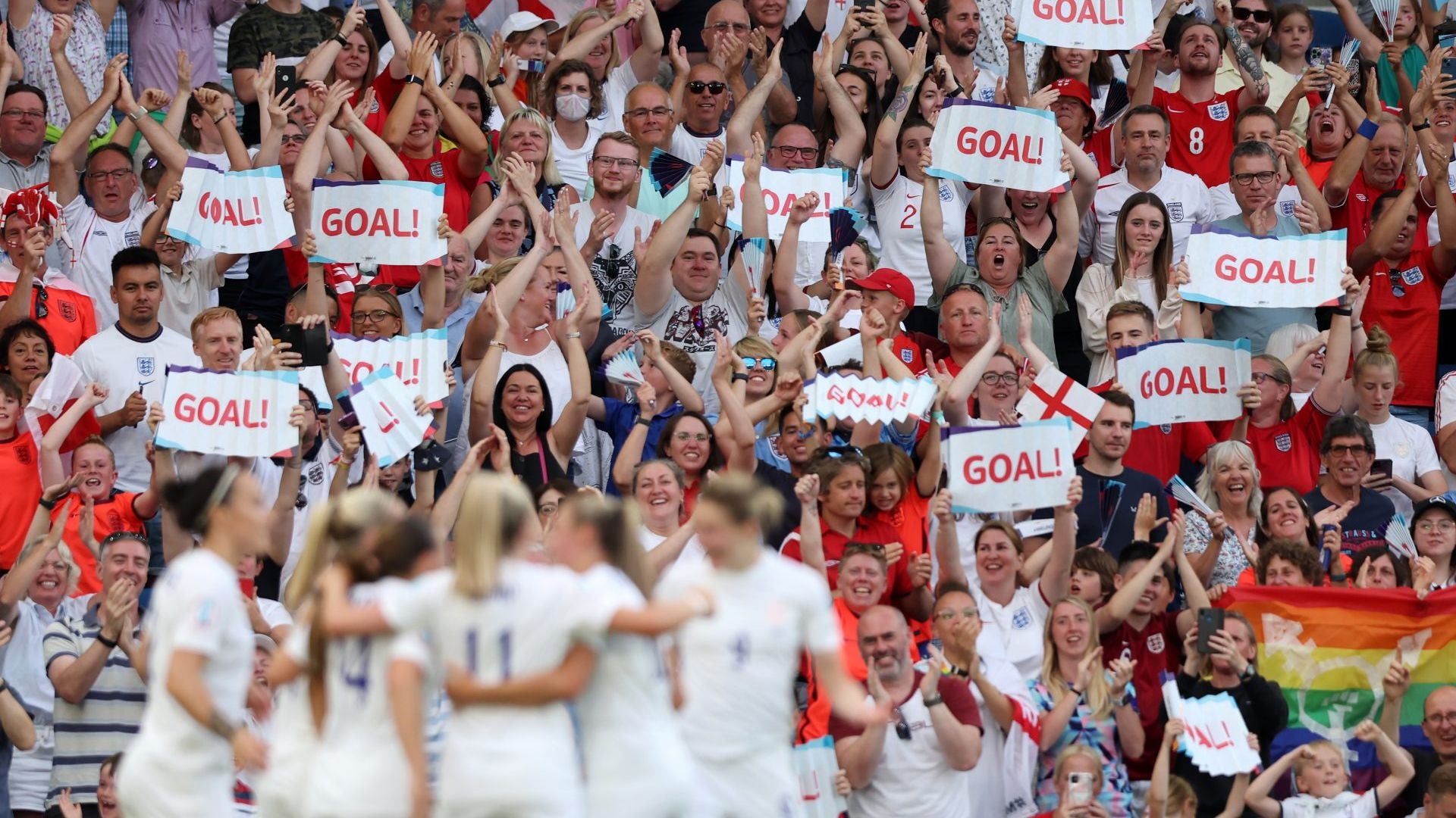 Fans in a stadium with 'GOAL!' signs celebrating, with players in white uniforms.
